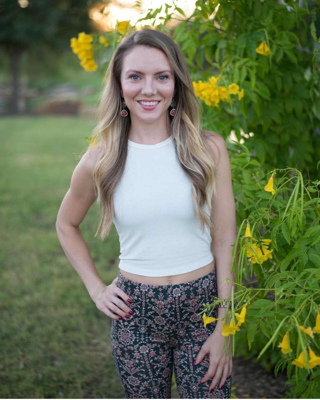 Woman in white top and floral pants beside yellow flowers.