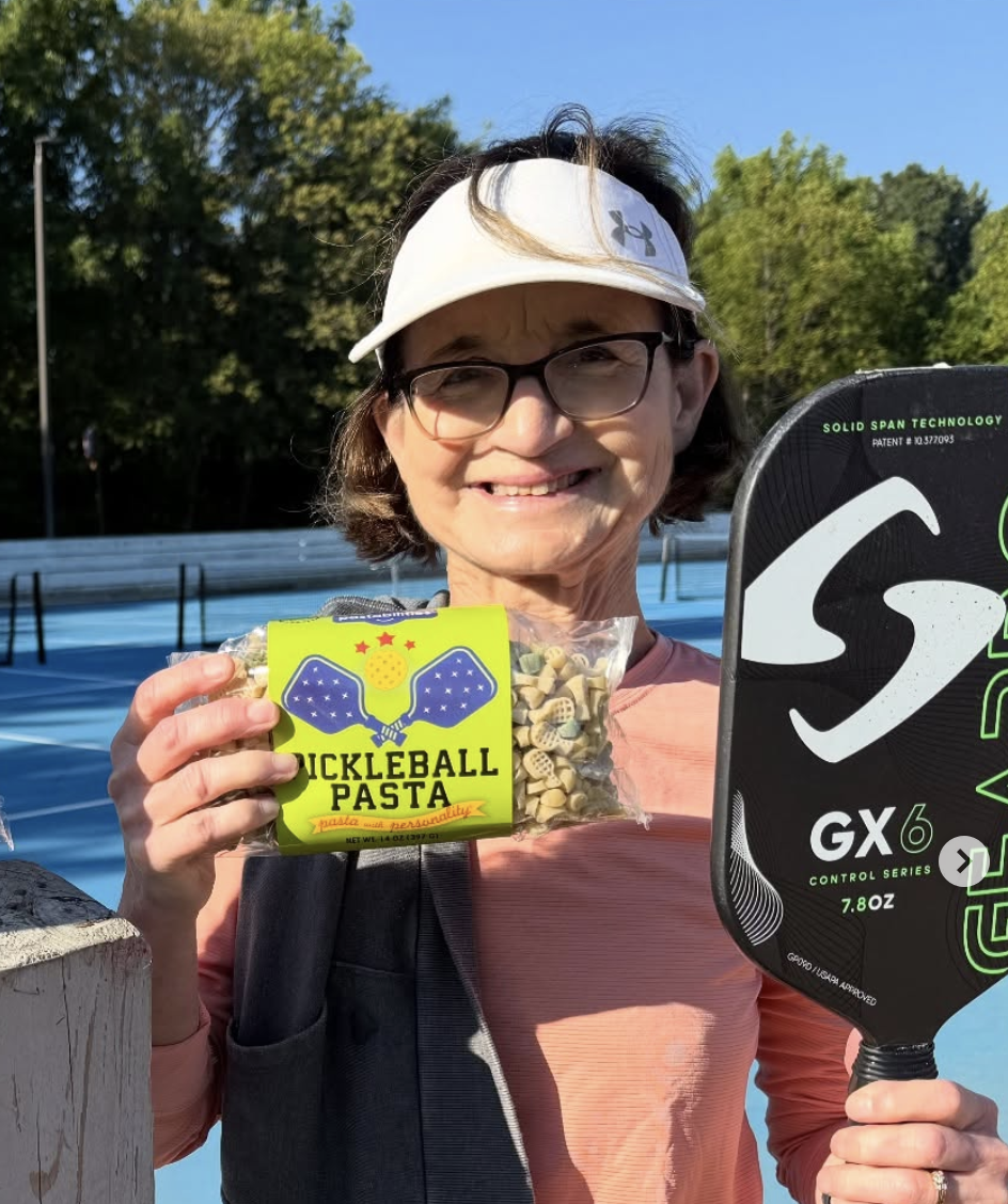 Smiling woman holding a bag of Pickleball Pasta and a pickleball paddle outdoors