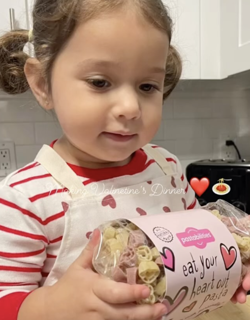 A young girl with pigtails in an apron holds a package of heart-shaped pasta.
