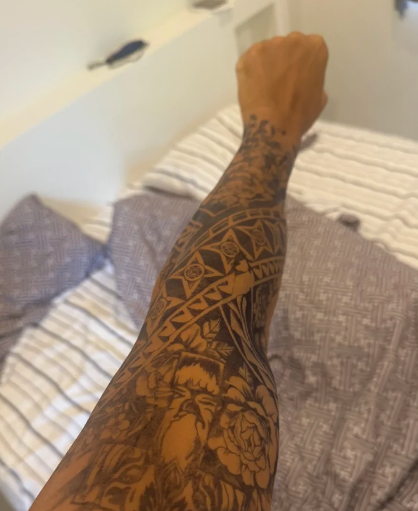 Arm with intricate floral and geometric tattoos, over a bed with patterned pillows.
