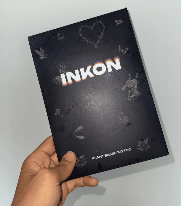 Hand holding a booklet titled 'INKON' with tattoo designs on the cover.