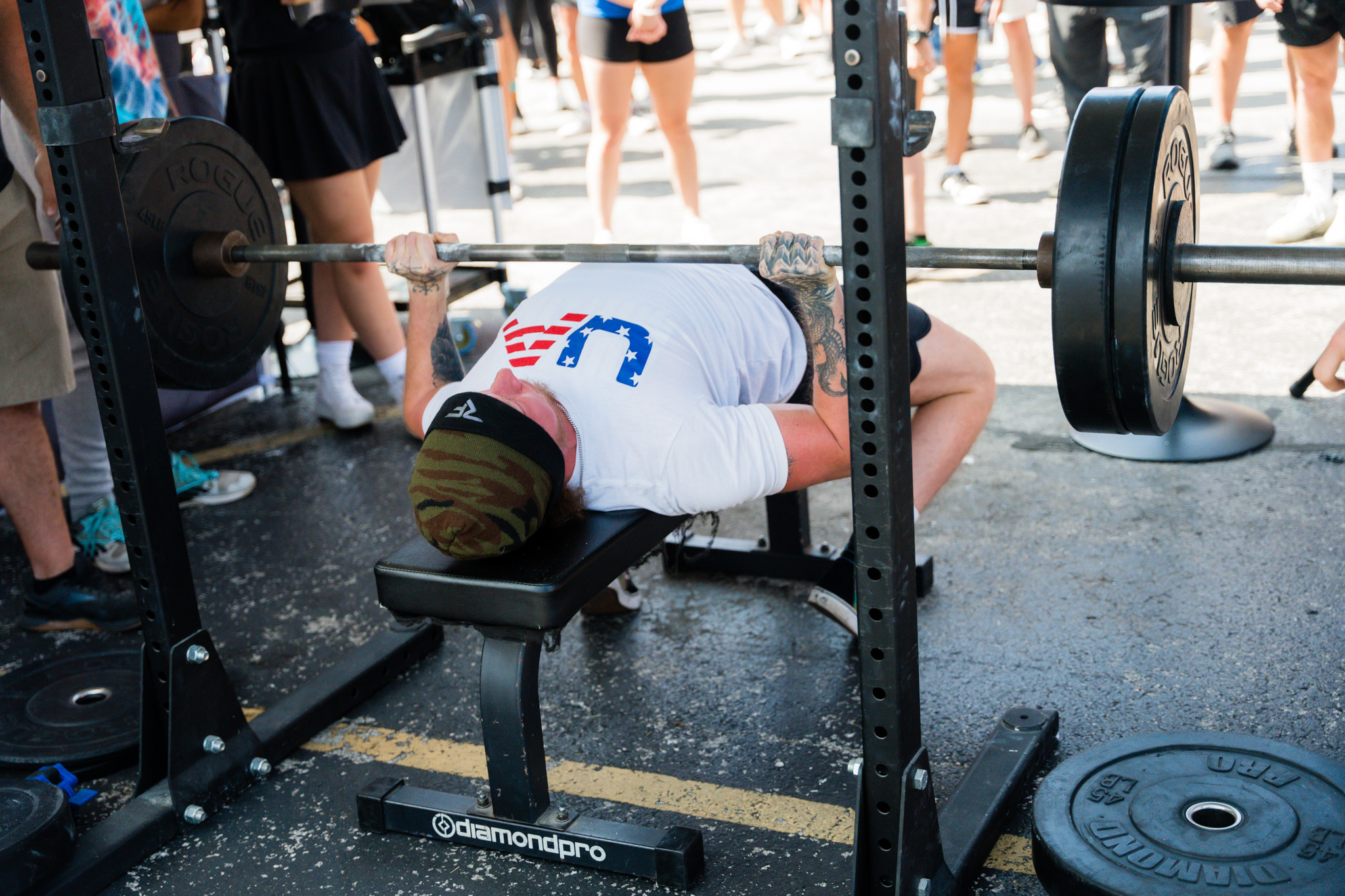 Person bench pressing weights outdoors with a crowd watching.