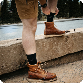 Person adjusting socks and leather boots on a roadside curb.