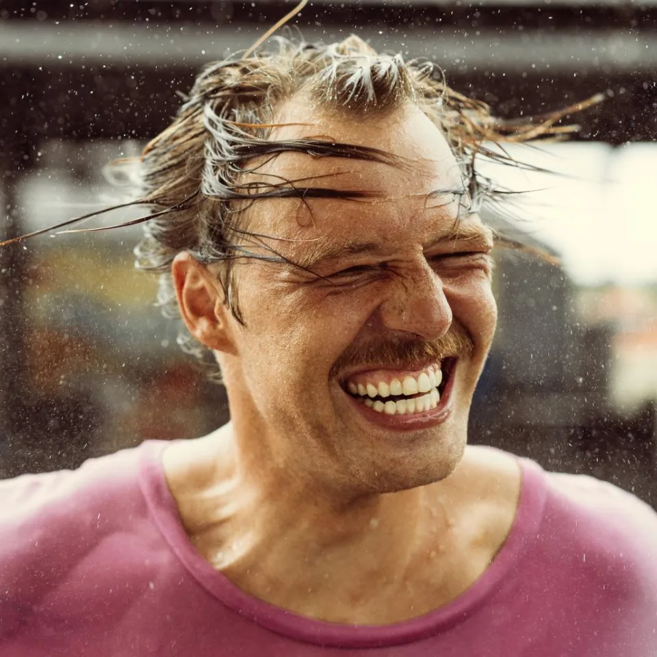 A man with a mustache laughs joyfully with his eyes shut as water sprays over his wet hair.