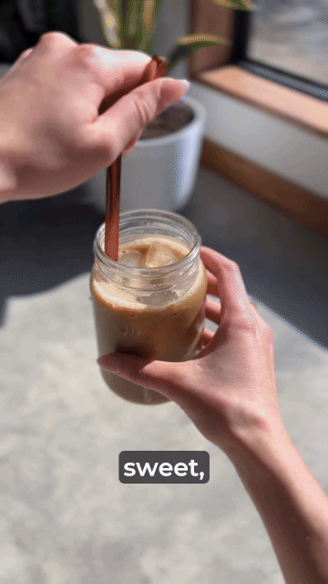 Hands stirring iced coffee in a jar with a wooden stick.