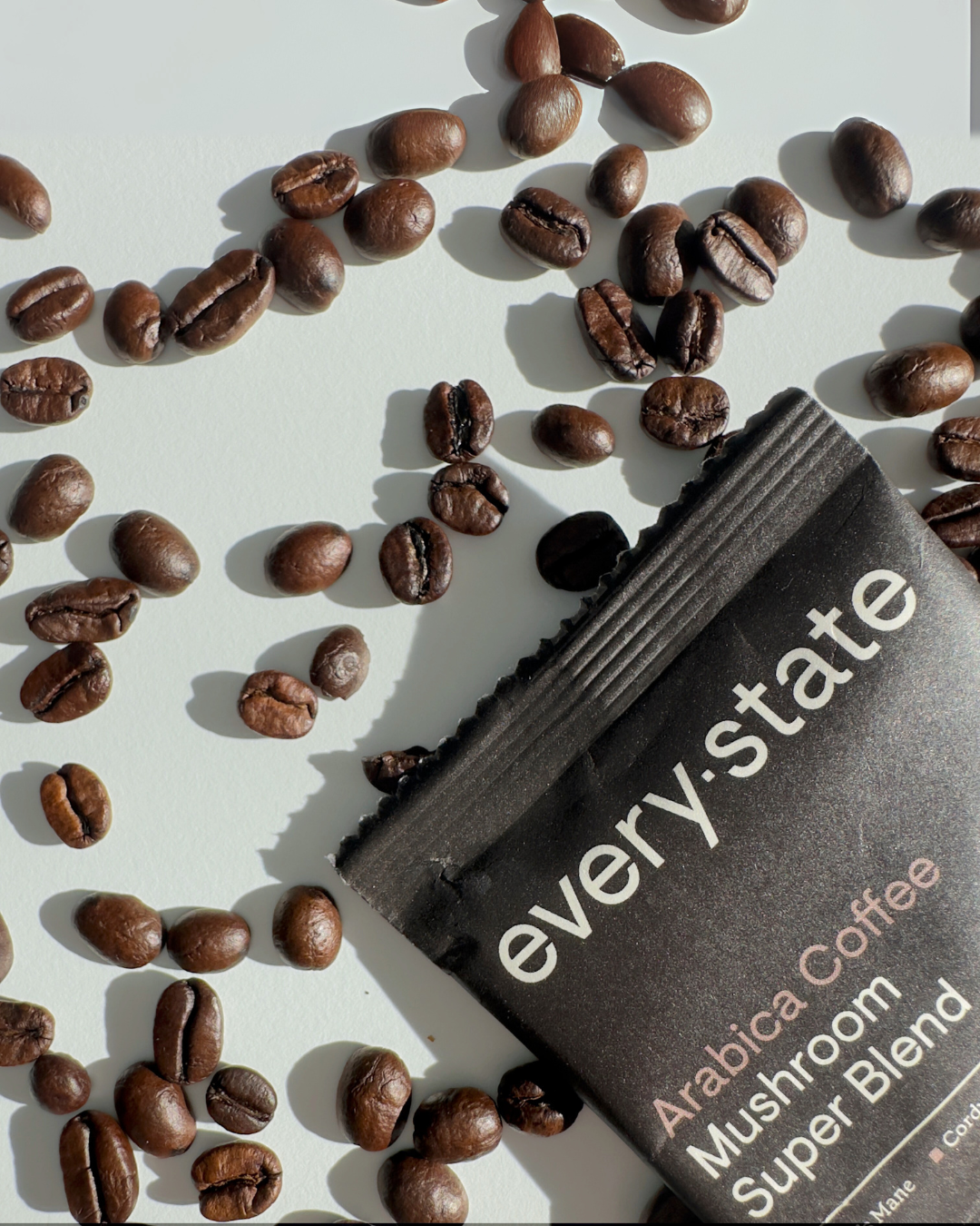 Scattered coffee beans with a packet labeled 'every.state Arabica Coffee Mushroom Super Blend.'