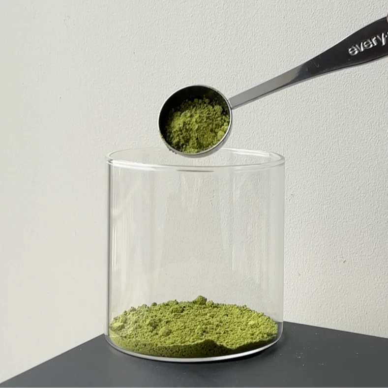 Spoon pouring green powder into a glass container on a dark surface.