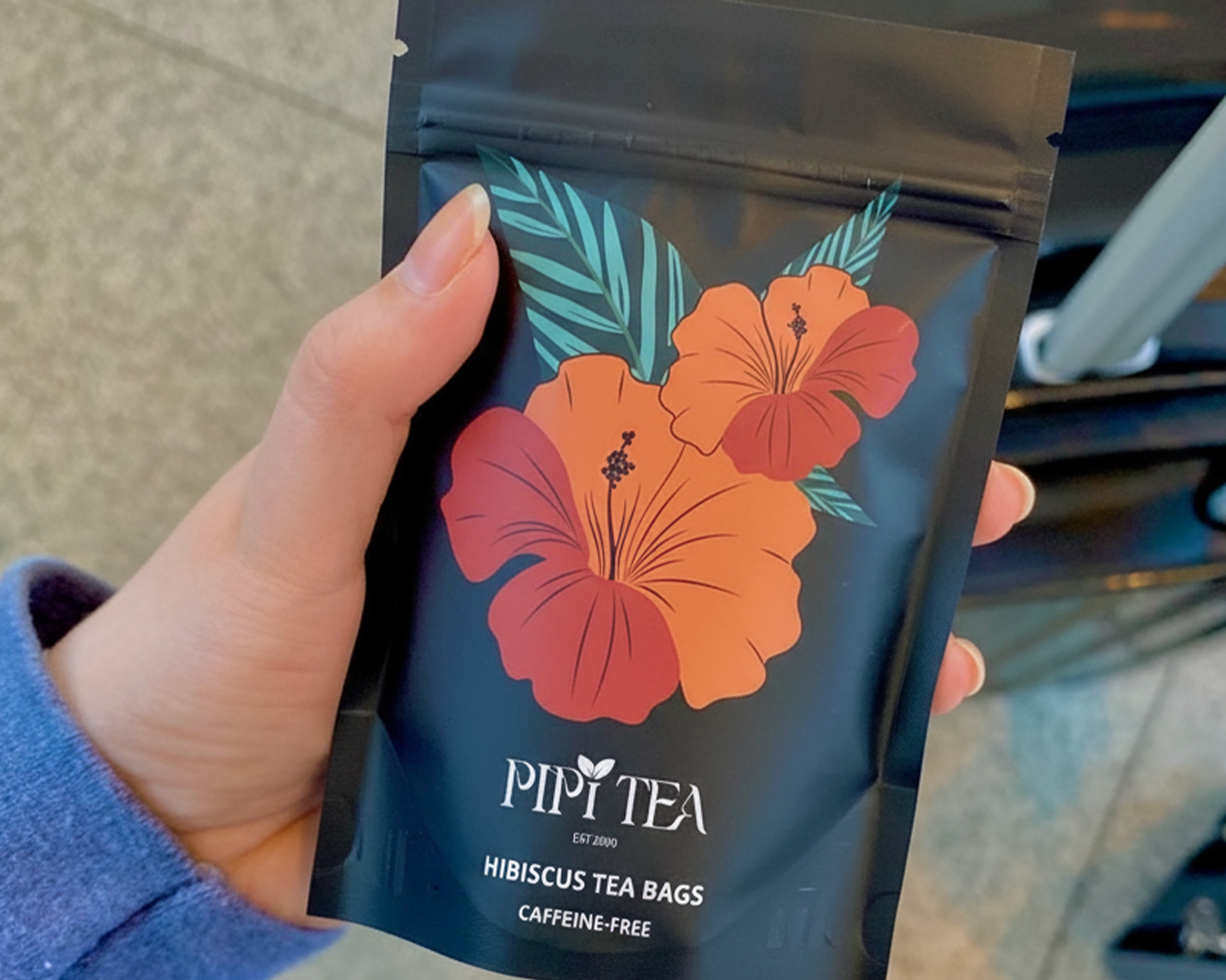 A person's hand holding a black pouch of Pipi Tea brand hibiscus tea bags.