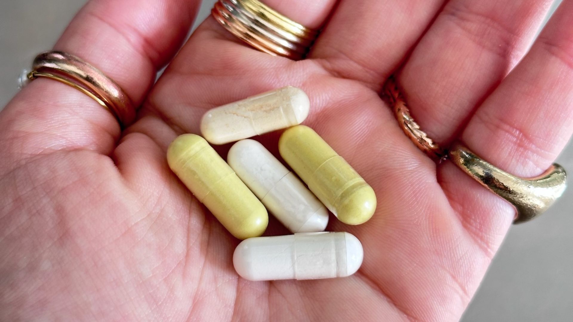 A close-up of a hand wearing several gold rings, holding five supplement capsules of various colors.