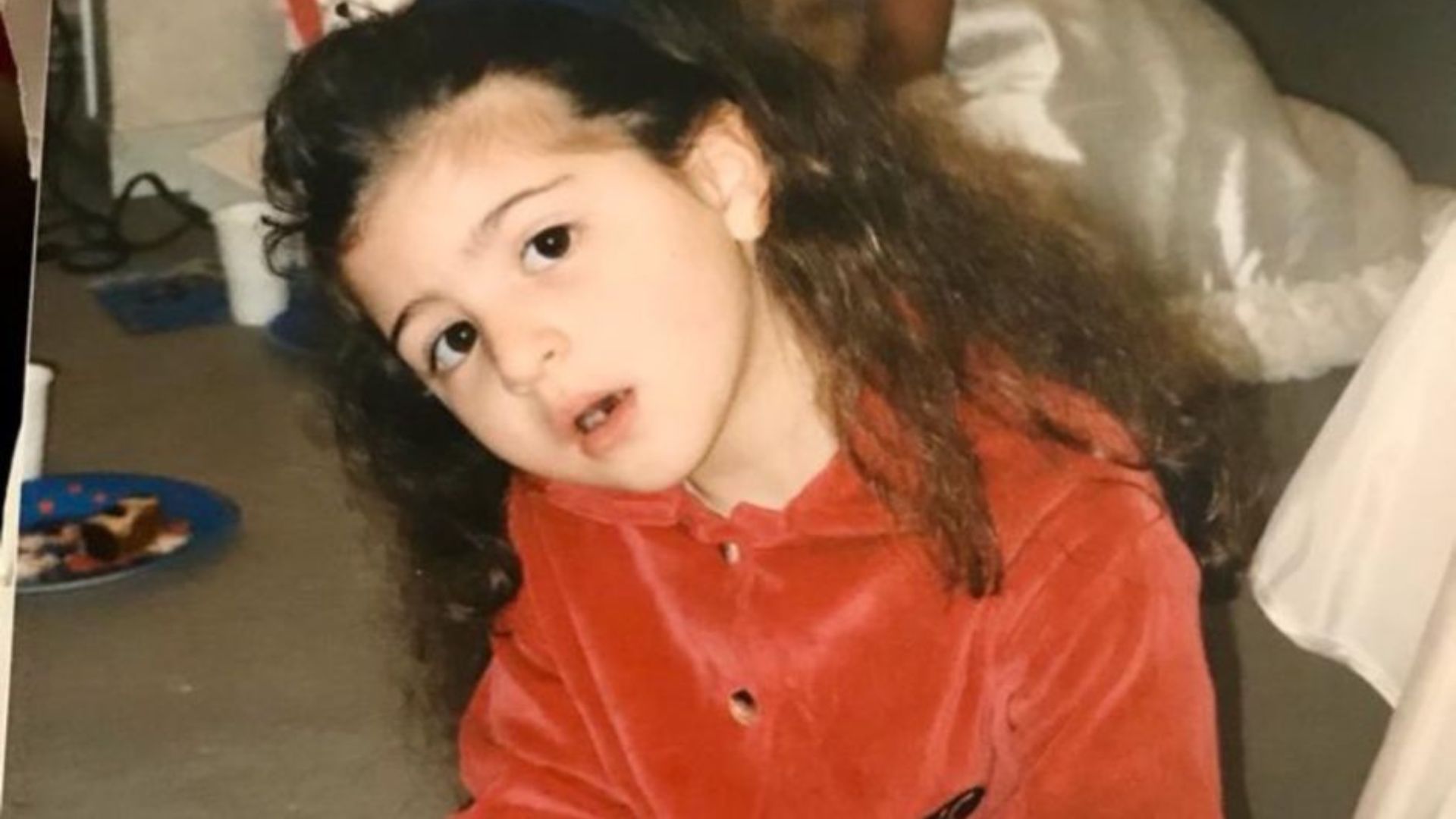A young girl with long, dark hair wearing a red collared shirt looks off-camera with her head tilted.
