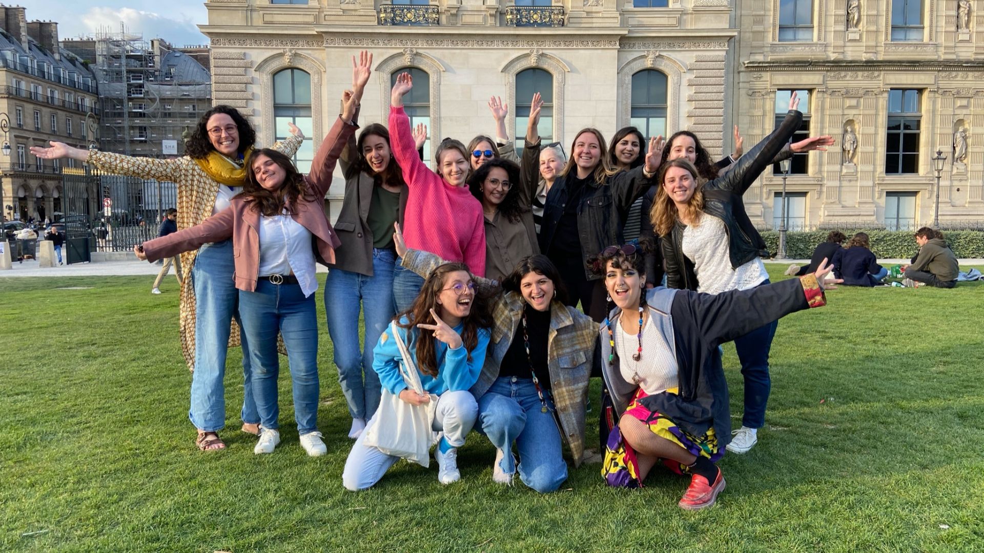 A group of women happily posing on a grassy lawn in front of a large, ornate building.