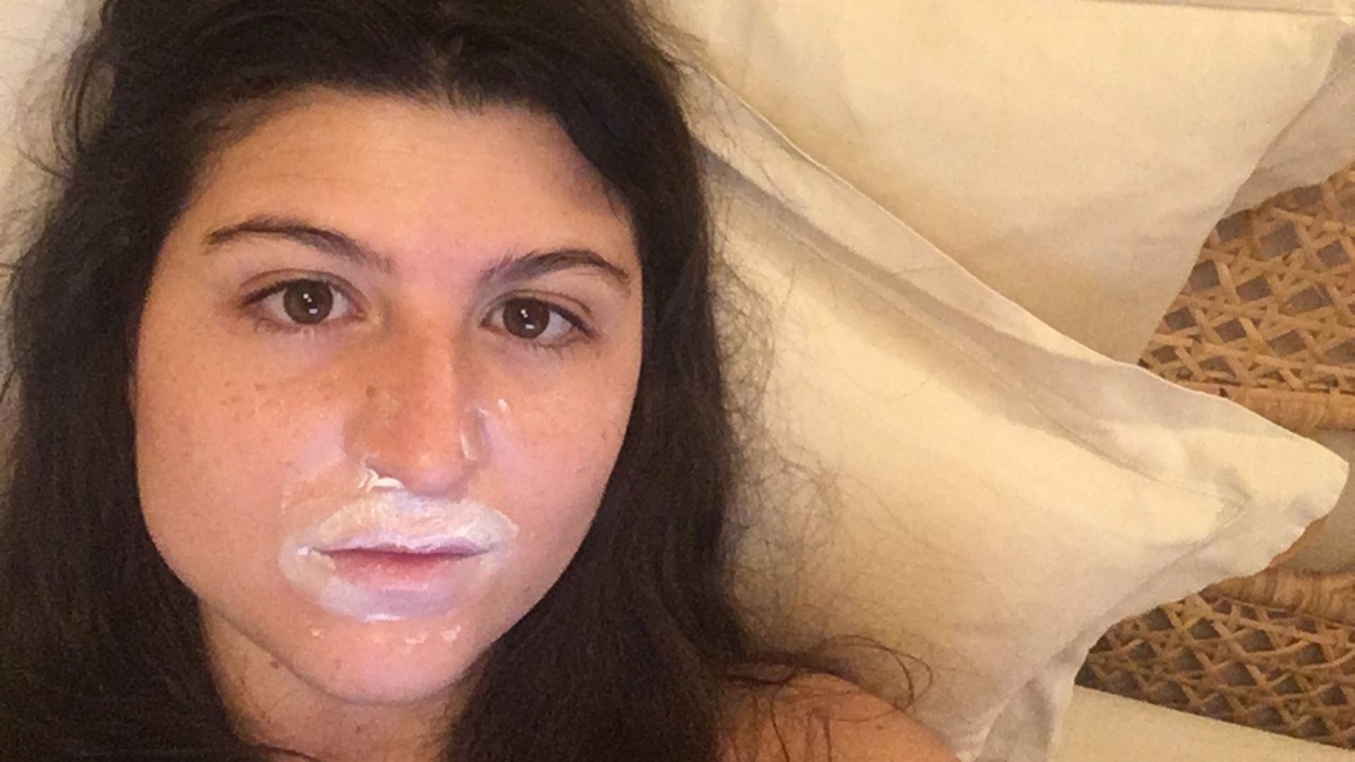 A close-up of a woman with long dark hair lying down with a white cream around her mouth.