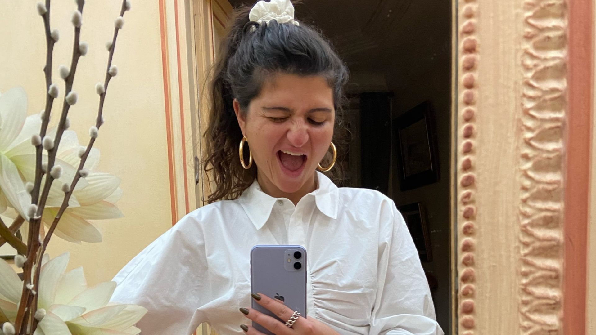A woman with her hair up takes a mirror selfie, making a silly, laughing face with eyes closed.