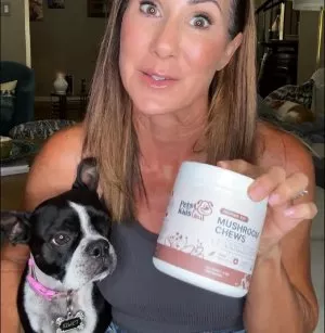A woman smiles while holding a dog in one arm and a jar of dog food in the other.