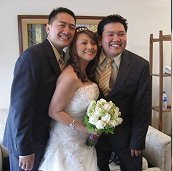 Three people in wedding attire, two men in suits, one woman in a white dress holding flowers.