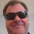 A close-up photo of a middle-aged man with brown hair wearing black sunglasses and smiling slightly.