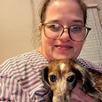 A woman with glasses and a striped shirt smiling while holding a small dog in front of her.