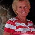 A smiling older woman with short, curly blonde hair, wearing a red and white striped shirt.