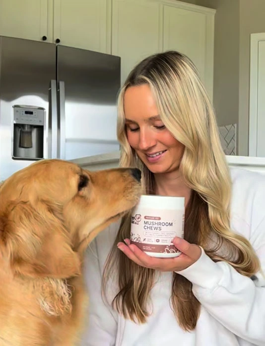 Woman holding mushroom chews container, smiling at a golden retriever in a kitchen.