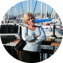 A blonde woman in a light blue sweater stands with her arms crossed in front of a boat marina.