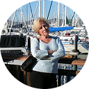 A woman with blonde hair and a light blue sweater stands with her arms crossed in front of sailboats.