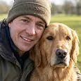 A smiling man in a beanie hat poses for a photo with his golden retriever dog in a field.