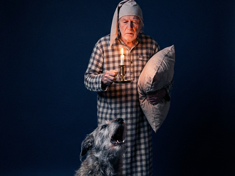 An elderly man in a nightgown and cap holds a candle and a pillow next to a howling dog.