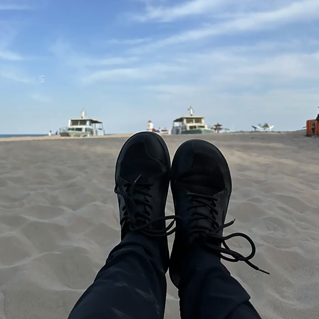Person's feet in black shoes on sandy beach, boats in background.