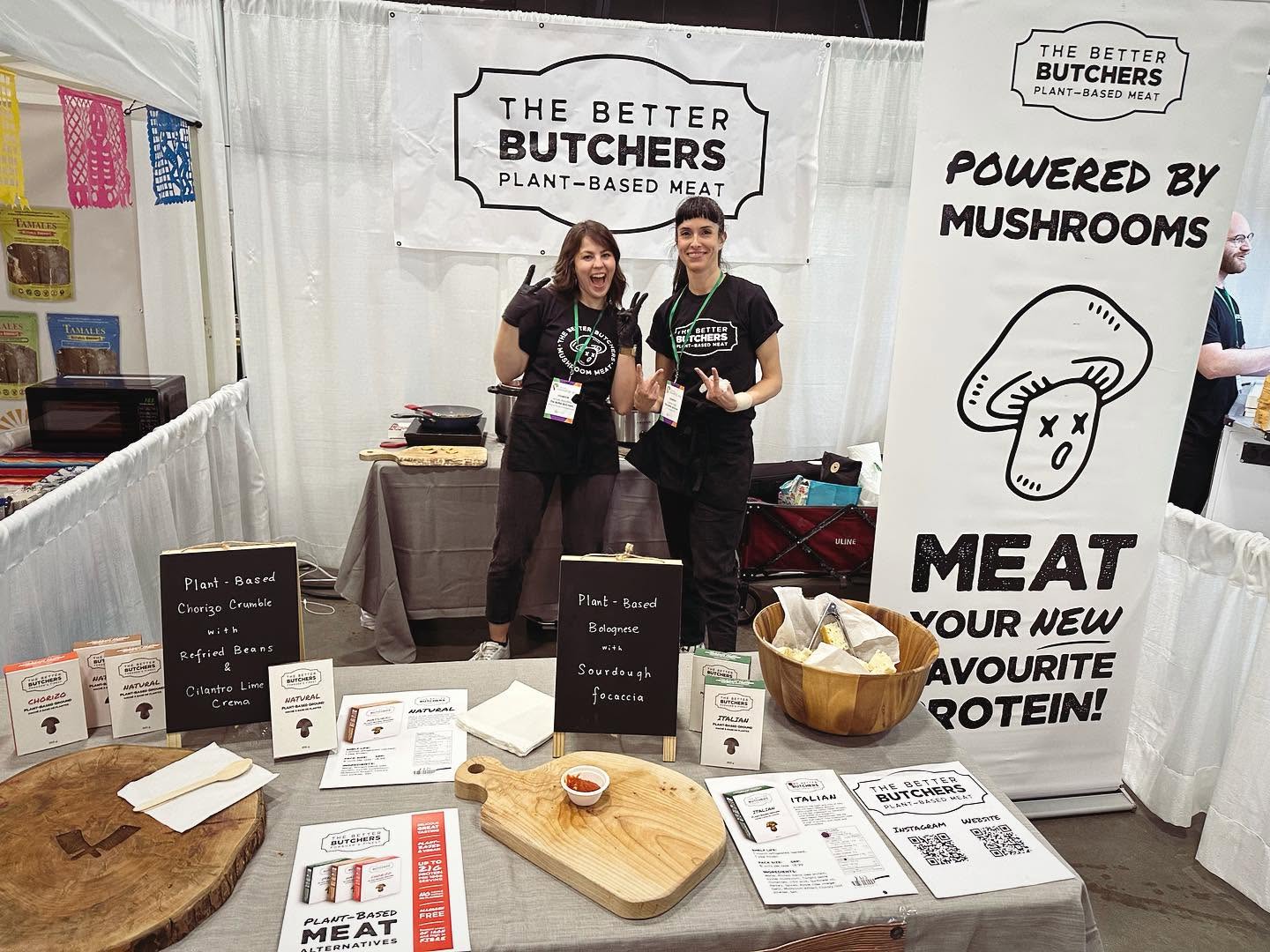 Two women stand at 'The Better Butchers' plant-based meat booth during a food expo, smiling for the camera.