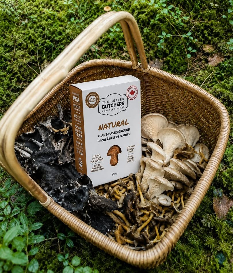 A wicker basket on moss is filled with various fresh mushrooms and a box of plant-based ground.