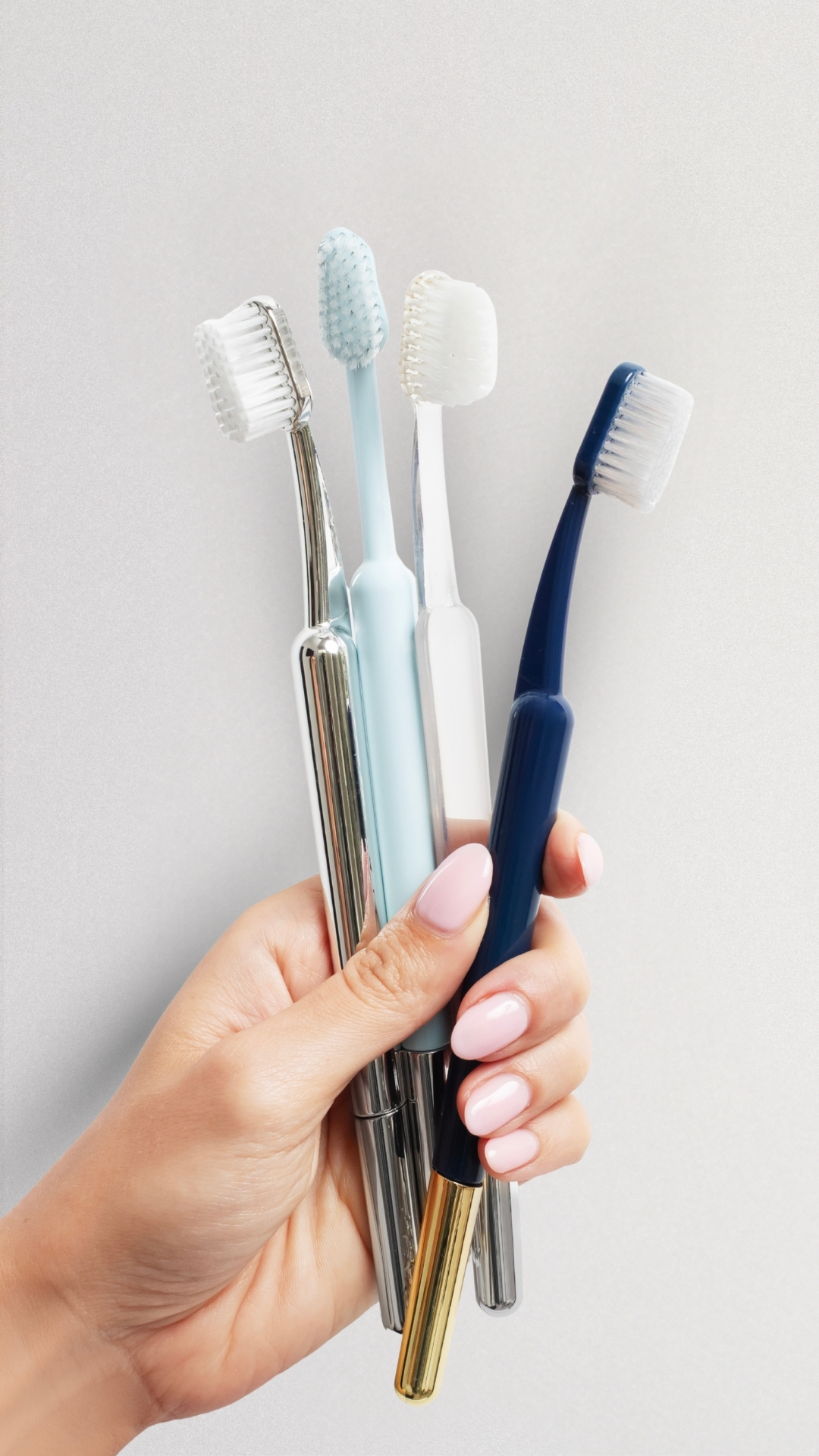 A hand with a light pink manicure holding a variety of five different toothbrushes.