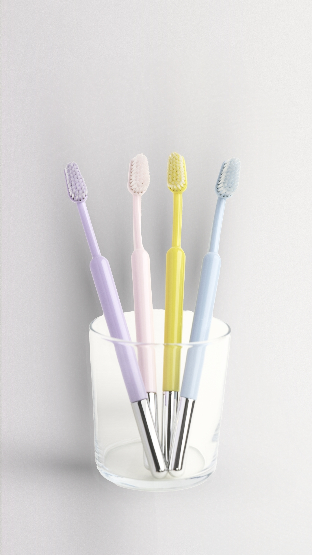 Four pastel-colored toothbrushes with silver-tipped handles standing in a clear glass against a white background.