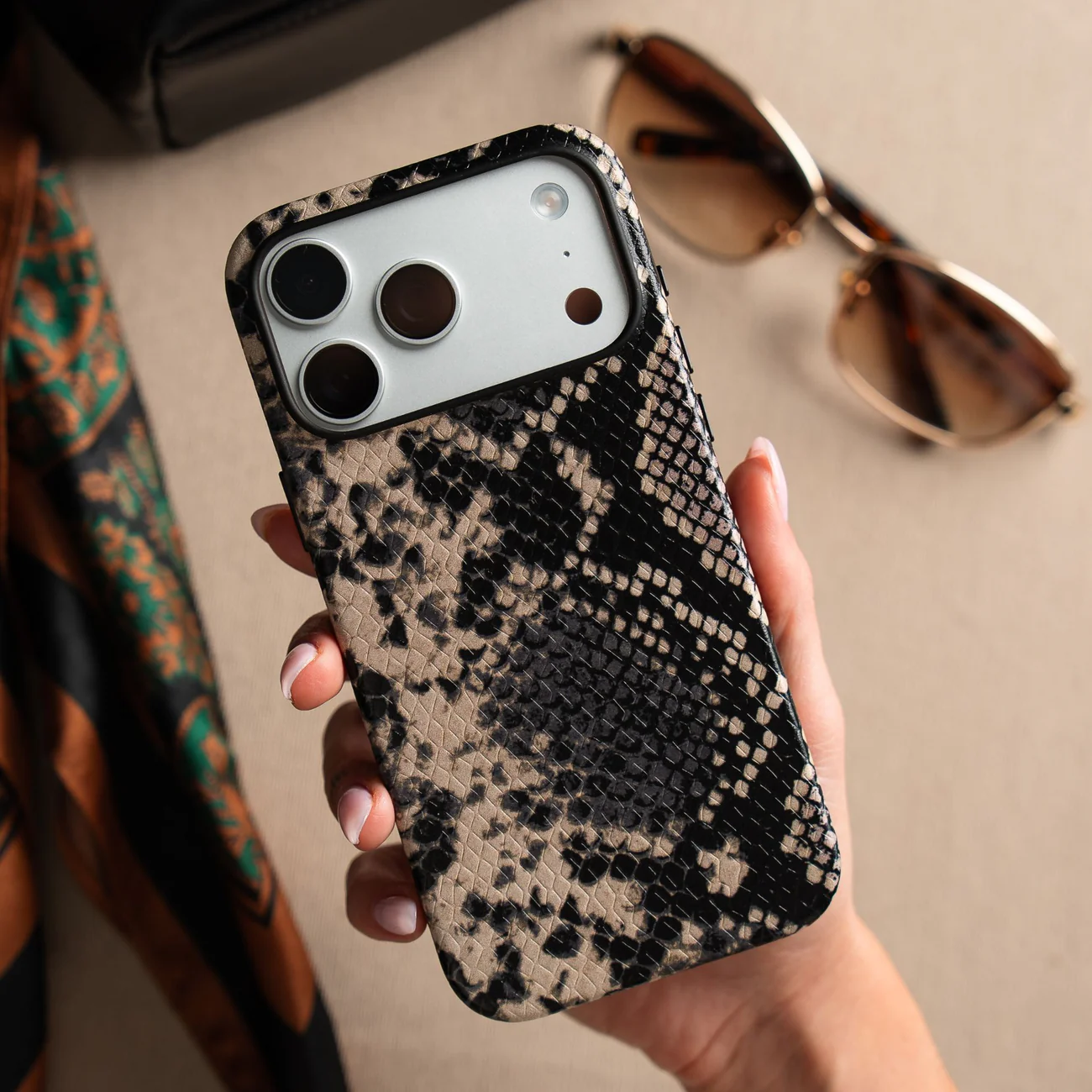 Hand holding phone with snake-patterned case beside sunglasses and scarf.
