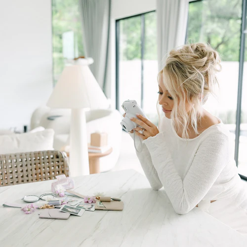 Woman sitting at a table with a smartphone, surrounded by photos and small items.