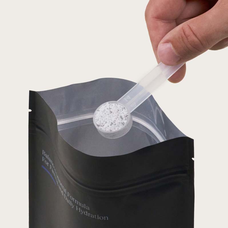 A hand holds a spoonful of powder over a black resealable pouch.