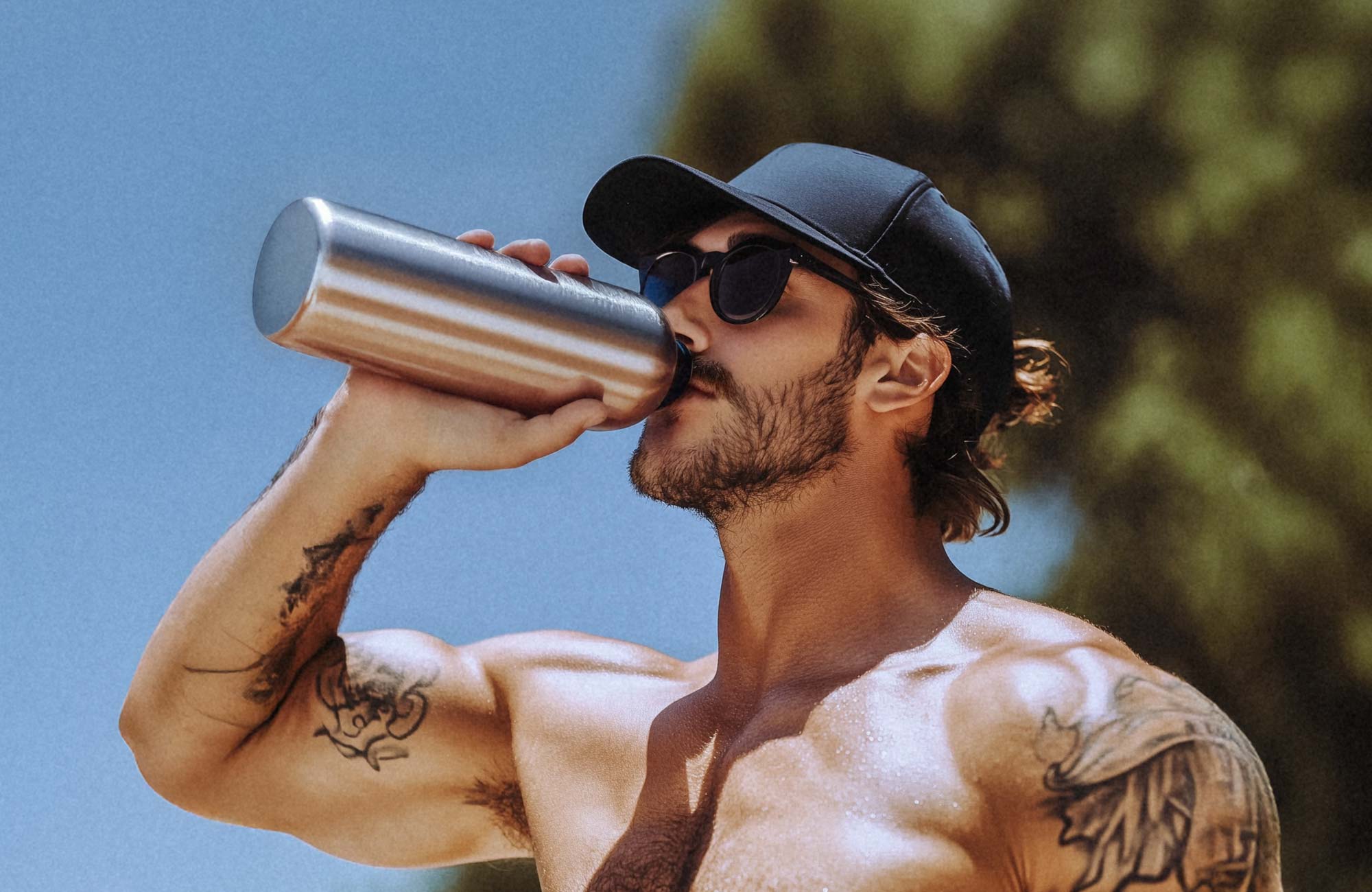 Man with tattoos drinking from a metal bottle outdoors.