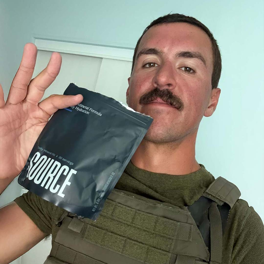 Person holding a black package labeled 'SOURCE' with a mustache and wearing a tactical vest.