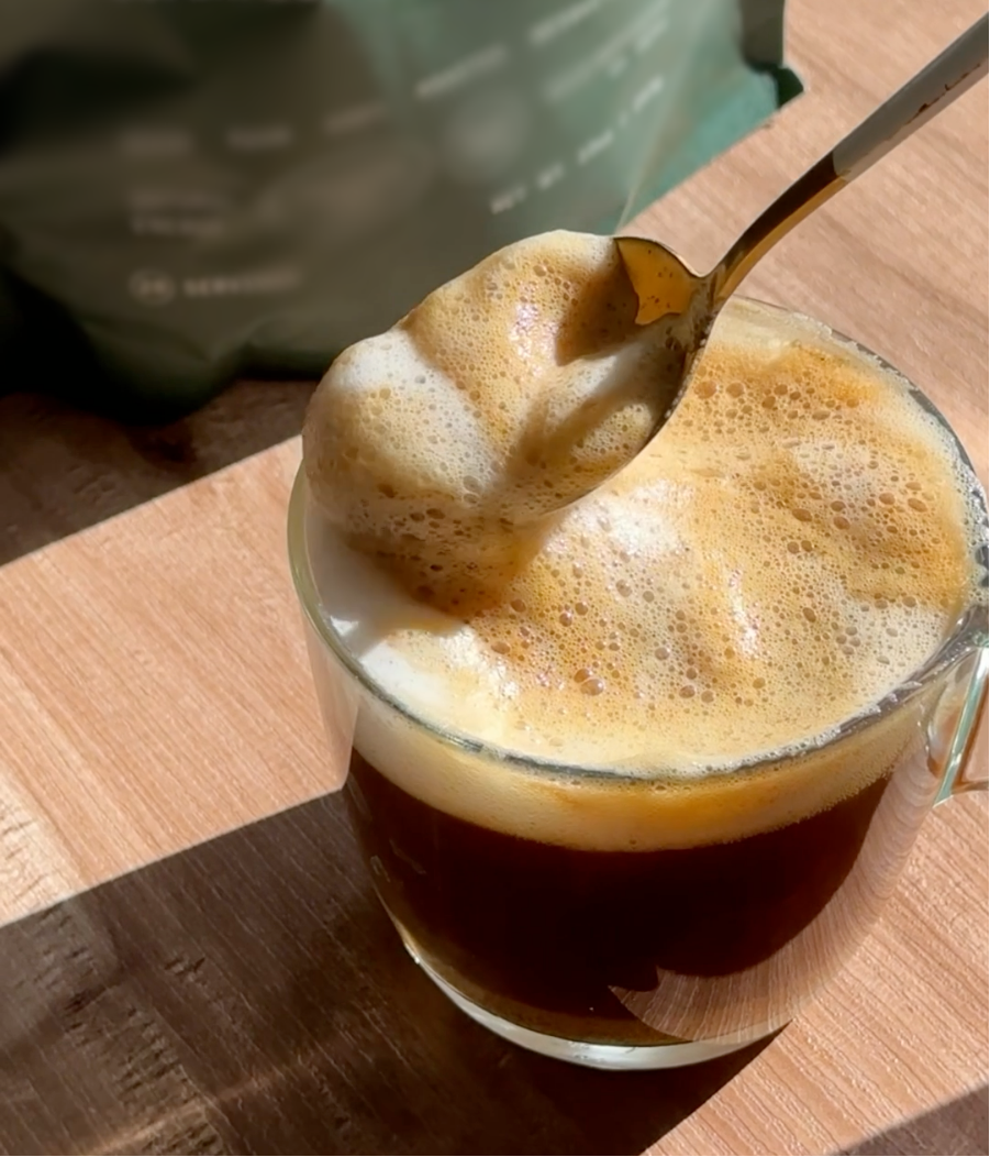 Glass cup of coffee with frothy milk and a spoon on a wooden table.
