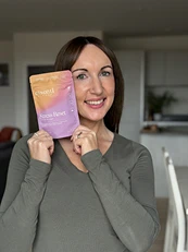 A smiling woman with brown hair holds a colorful product pouch next to her face.