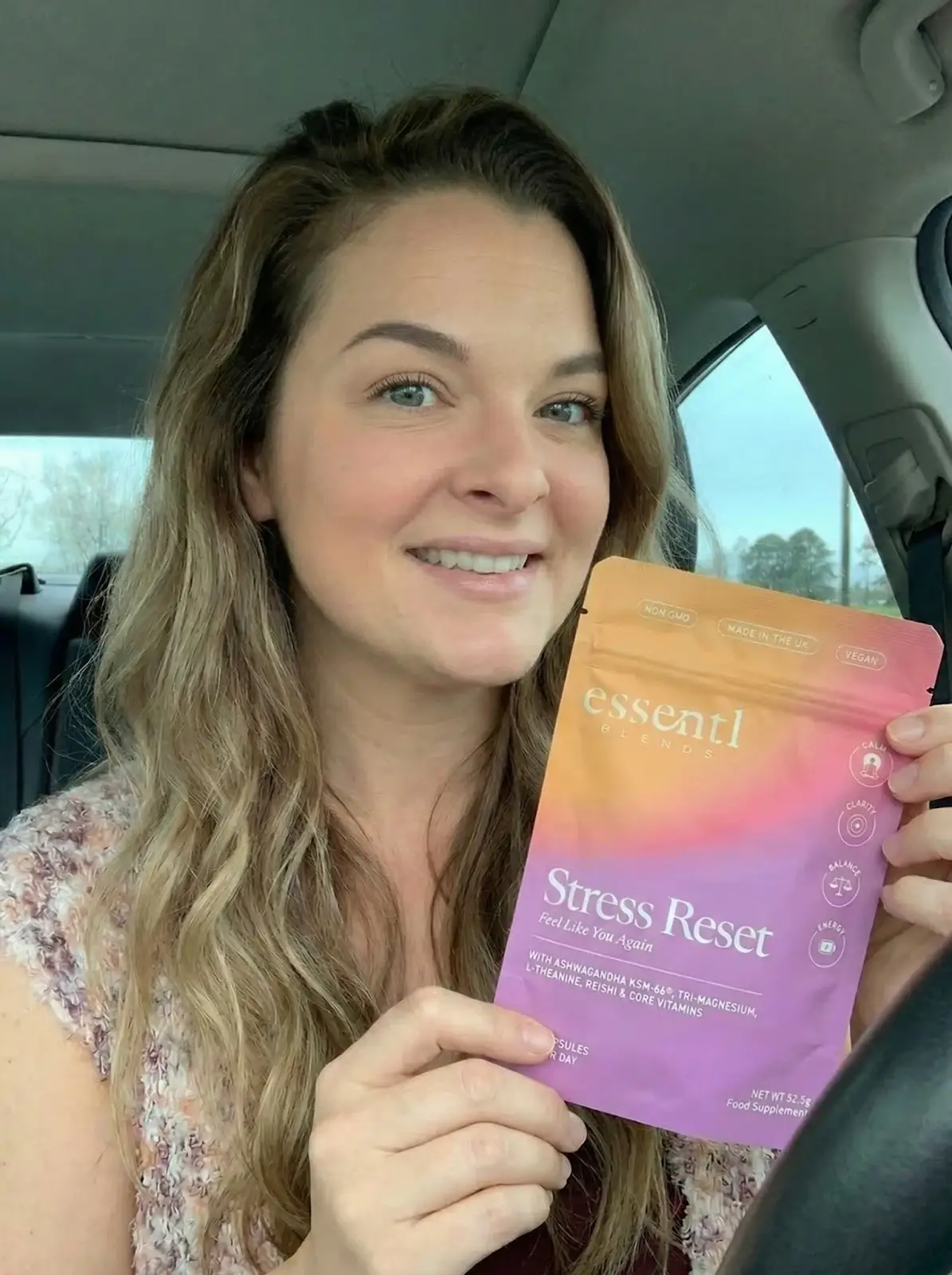 A smiling woman inside a car holds up a purple and orange pouch of essentl BLENDS Stress Reset.