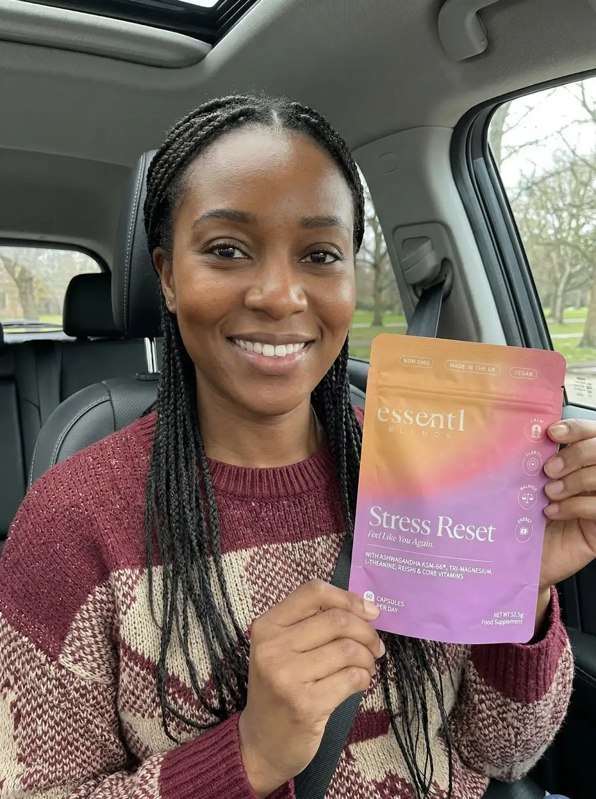 A smiling woman with braided hair sits in a car, holding a pouch of Essentl Blends Stress Reset.