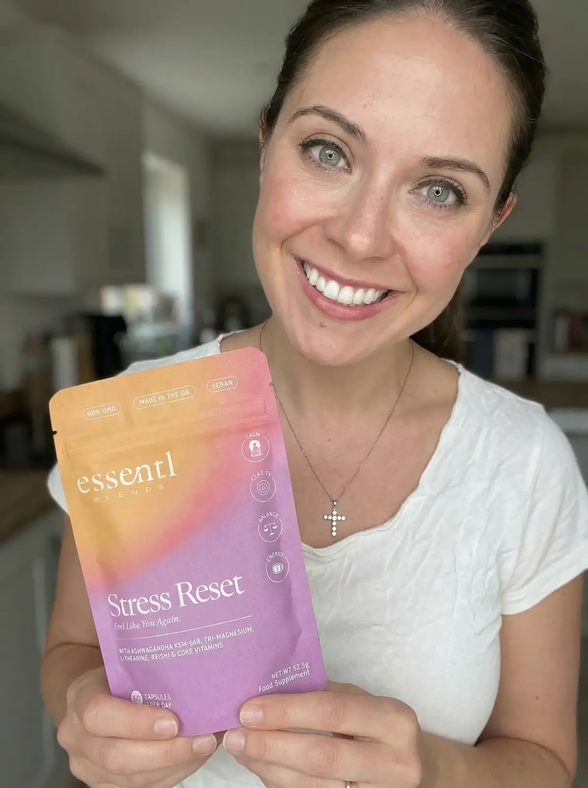 A smiling woman in a white shirt holds up a pouch of Essentl Blends Stress Reset supplements.