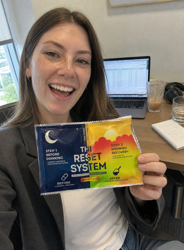 A smiling woman at a desk holds up a packet of 'The Reset System' supplement.