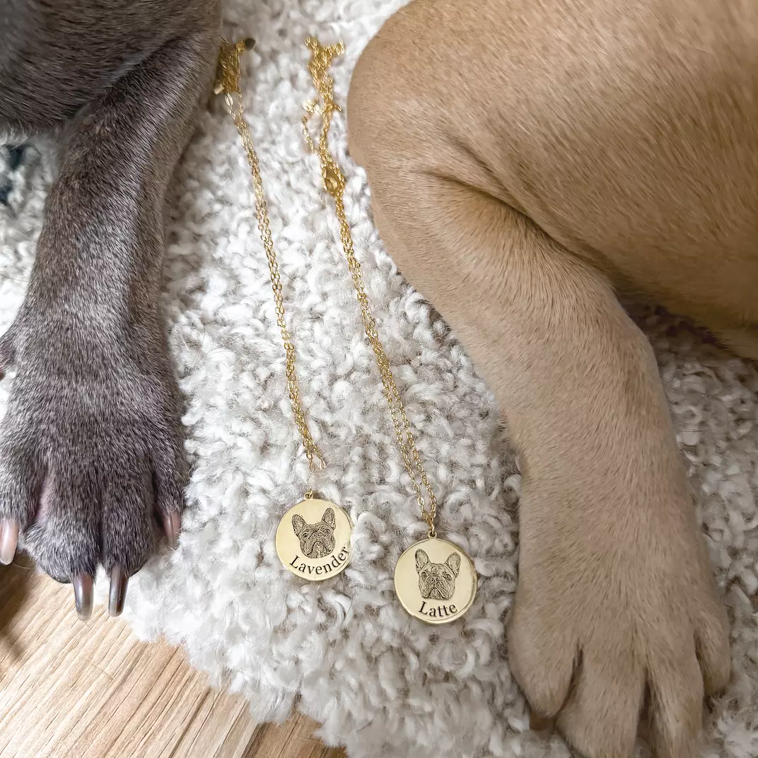 Two gold pendant necklaces rest on a fluffy white rug next to the paws of two dogs. The dogs appear to be a gray French Bulldog on the left and a tan French Bulldog on the right. 