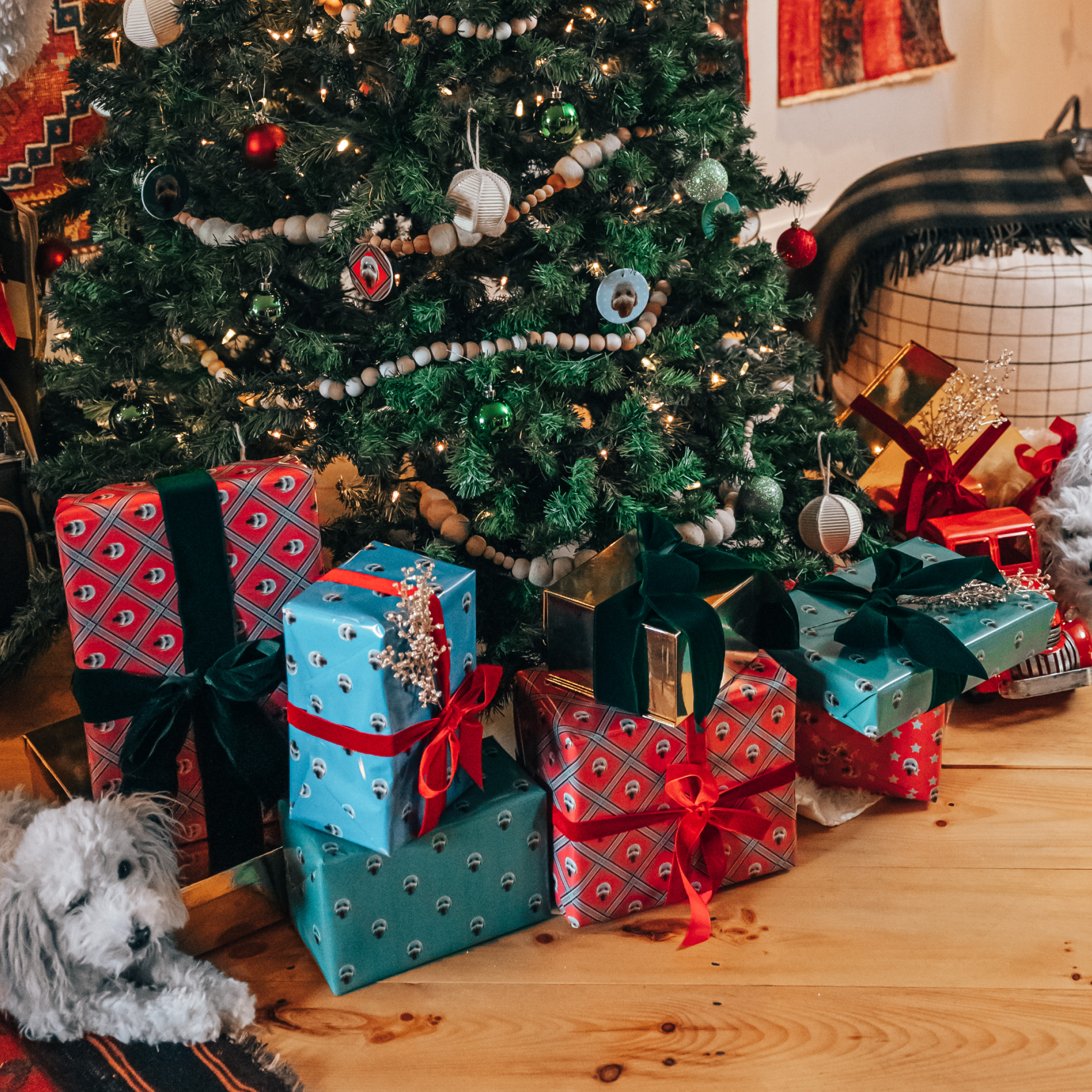 A silver-gray plush replica of a fluffy dog lies on a rug beneath a decorated Christmas tree surrounded by wrapped presents in red and blue patterned paper.