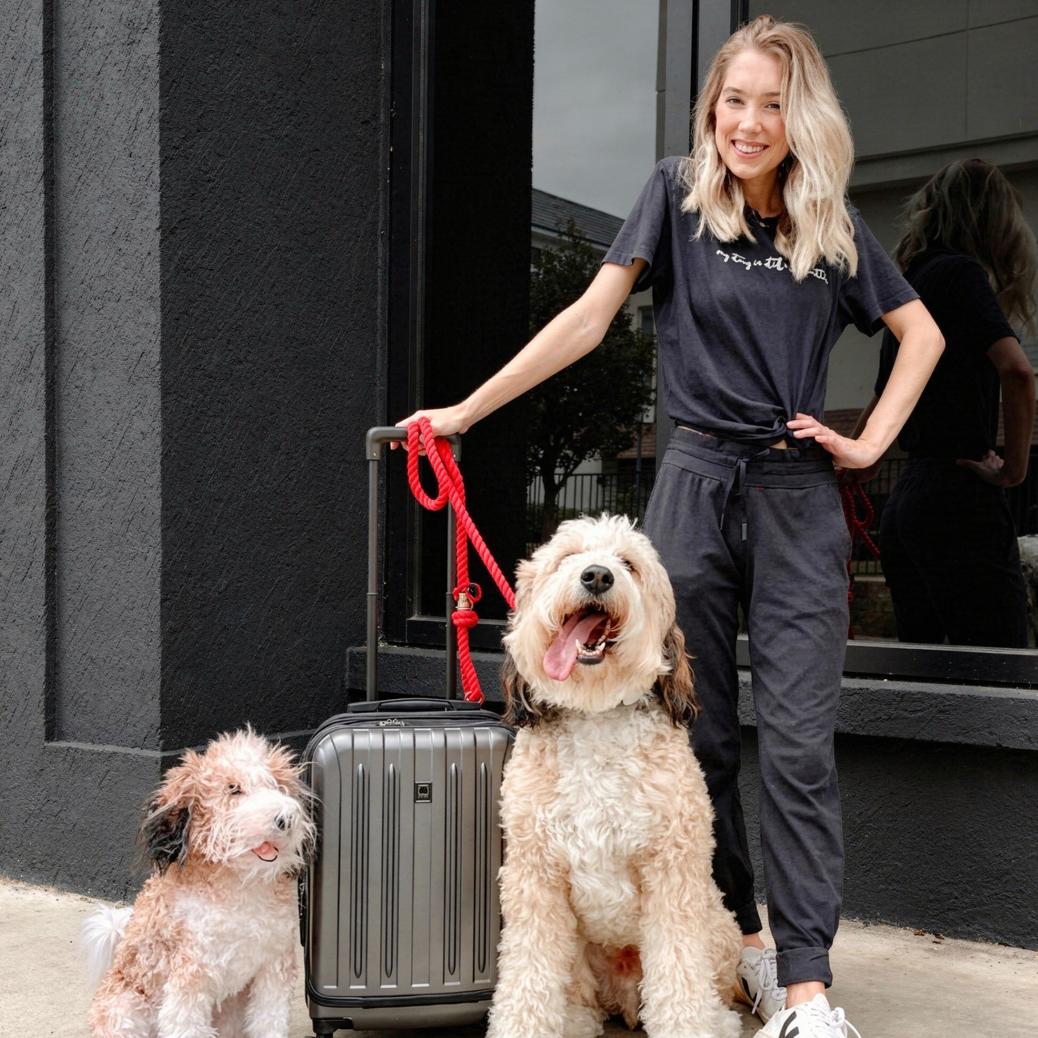 Smiling woman stands outside with silver suitcase, real & plush fluffy dogs.