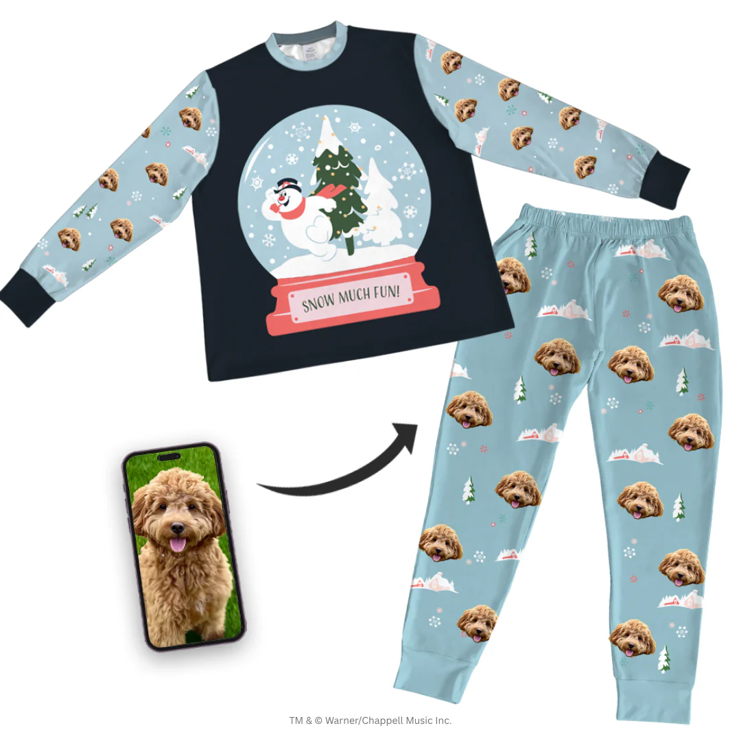 A dark blue, long-sleeve pajama top features a large graphic of Frosty the Snowman and a tree inside a snow globe that reads 'SNOW MUCH FUN!' The sleeves are light blue and patterned with a fluffy brown Cockapoo's face, small trees, and snowflakes. The matching light blue pajama pants are patterned with the Cockapoo's face and snow scene graphics.
