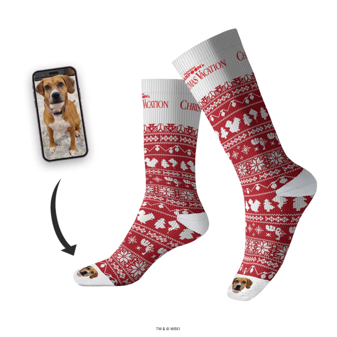 A pair of tall white socks with a red and white fair isle pattern featuring Christmas Vacation-themed silhouettes. The cuff of the socks is white with red text reading 'NATIONAL LAMPOON'S CHRISTMAS VACATION.' The toe of each sock features an image of a brown and white dog's face. A smartphone screen inset shows the original photo of the dog, with an arrow pointing to the socks.
