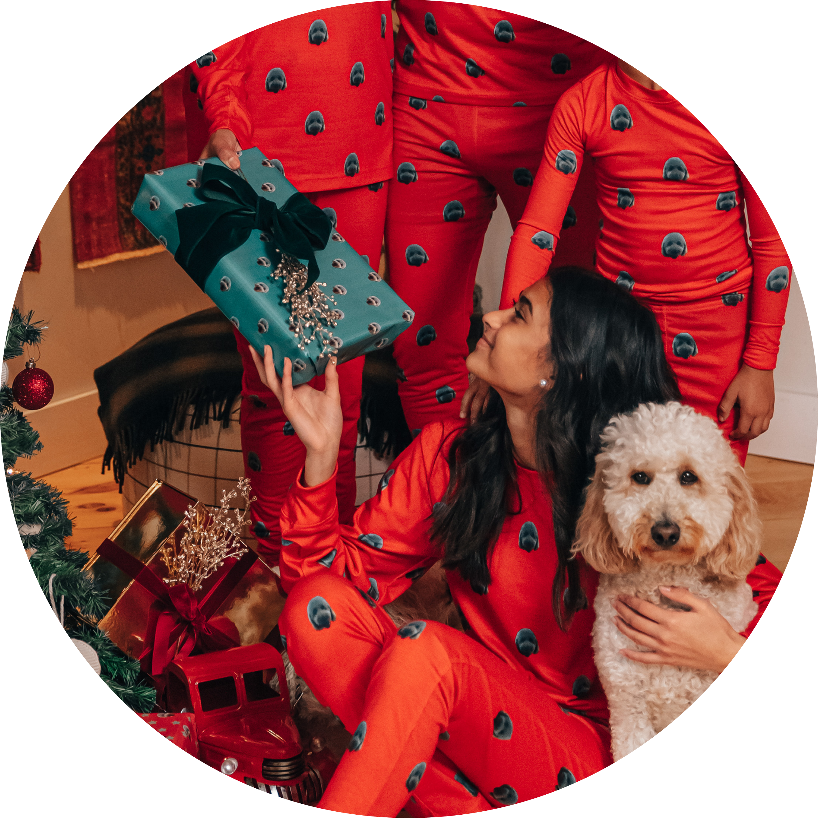 A young woman sits on the floor holding a fluffy light brown dog and a gift wrapped in teal paper, while other family members stand around her. All the people are wearing red pajamas patterned with a repeating dog face.