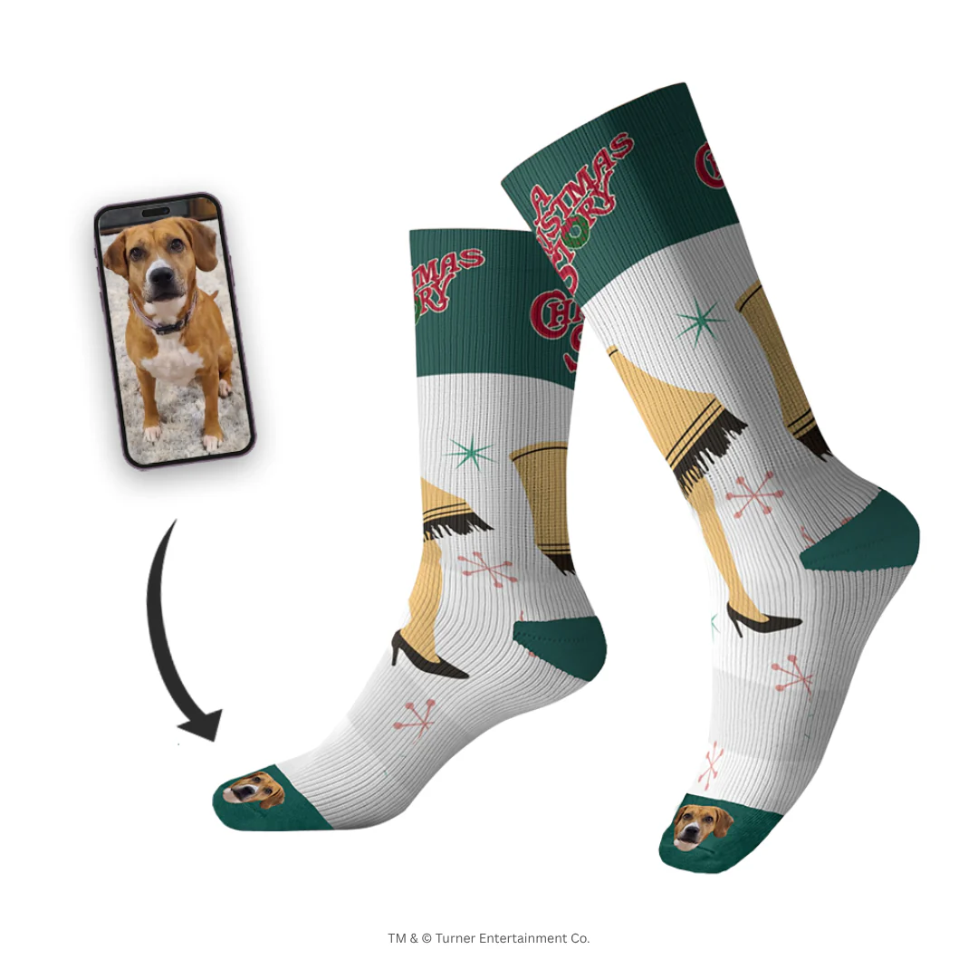 Promotional image for custom pet socks. A pair of tall white socks with green cuffs features graphics of the Leg Lamp from A Christmas Story and the movie title, interspersed with pink and white starbursts. The toe of each sock features an image of a brown and white dog's face. A smartphone screen inset shows the original photo of the dog, with an arrow pointing to the socks.